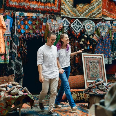 Two tourists walking through a vibrant market filled with colorful rugs, textiles, and traditional crafts. The surroundings are rich in cultural patterns and details, creating a lively and authentic atmosphere.