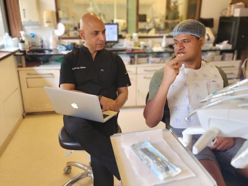 Doctor consulting a patient in a dental clinic, using a laptop while the patient gestures and discusses concerns