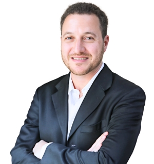 Professional headshot of Dr. Simon Flikier, smiling wearing a black suit and white shirt, standing with arms crossed against a white background.