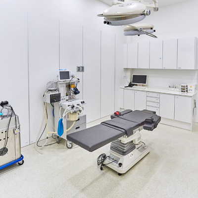 A state-of-the-art operating room with a sleek, modern design. The room features a centrally positioned black surgical table with adjustable sections, surrounded by high-tech medical equipment. Overhead, a large surgical light provides focused illumination. Against the white-paneled walls, a workstation with a computer and various medical supplies is neatly organized in white cabinetry. To the left, an anesthesia machine and other essential devices are mounted on mobile carts for easy maneuverability. The room's clean, sterile environment is complemented by a wooden door, adding a subtle contrast to the predominantly white and clinical aesthetic.