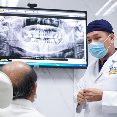 A dentist wearing a surgical mask and cap explains a dental X-ray to an elderly patient in a modern clinic. The X-ray image is displayed on a large screen.